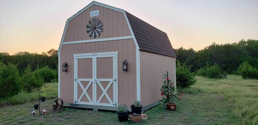 A tan barn-style shed with a windmill decoration on the front, surrounded by potted plants and garden tools, sits on a grassy field at sunset.