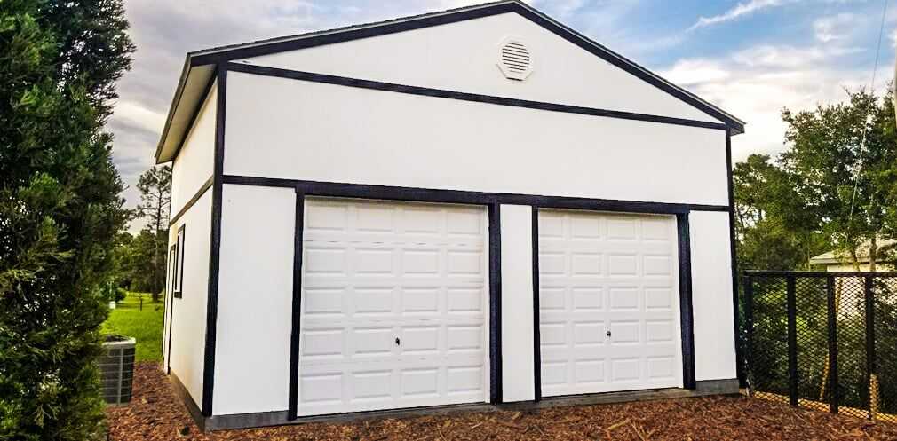 A white garage with two closed doors, black trim, and a vent at the top, surrounded by trees and a fenced area, under a partly cloudy sky.