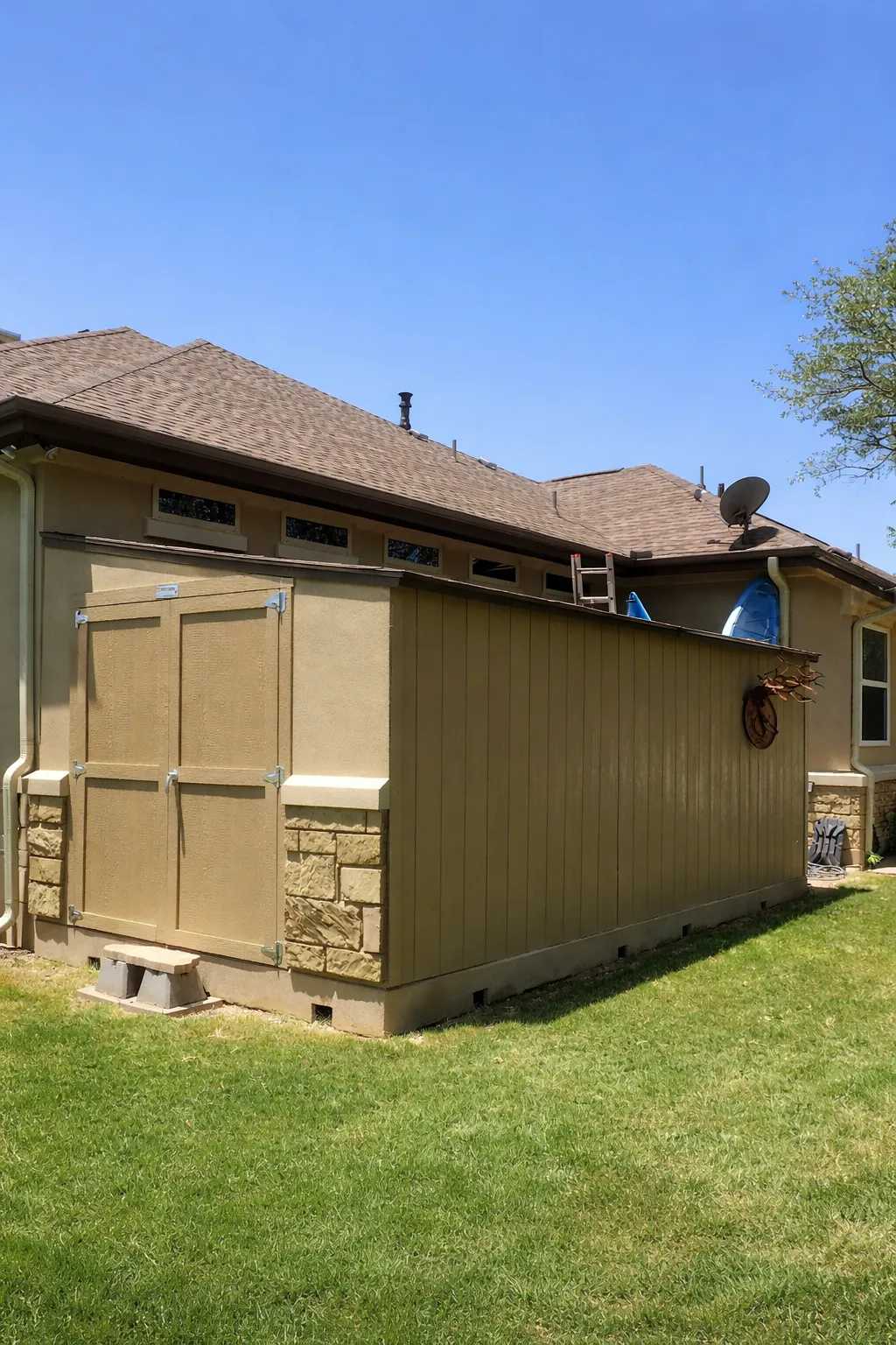 A light brown shed attached to the side of a house, with a ladder and wheelbarrow on top, set on a well-kept grassy lawn under a clear blue sky.
