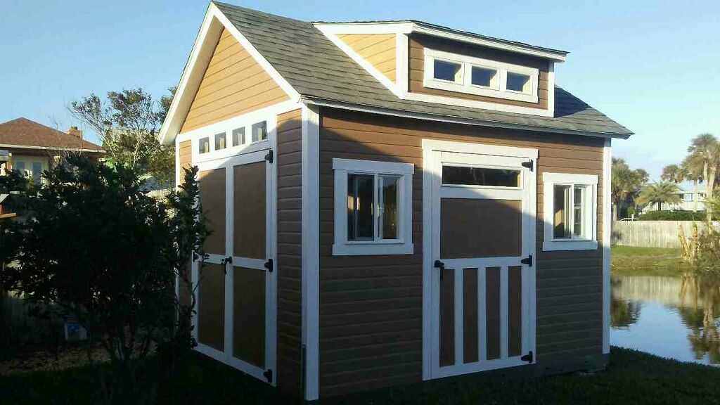 A small, brown and white Tuff Shed building with a gabled roof and multiple windows, situated near a pond and surrounded by greenery.