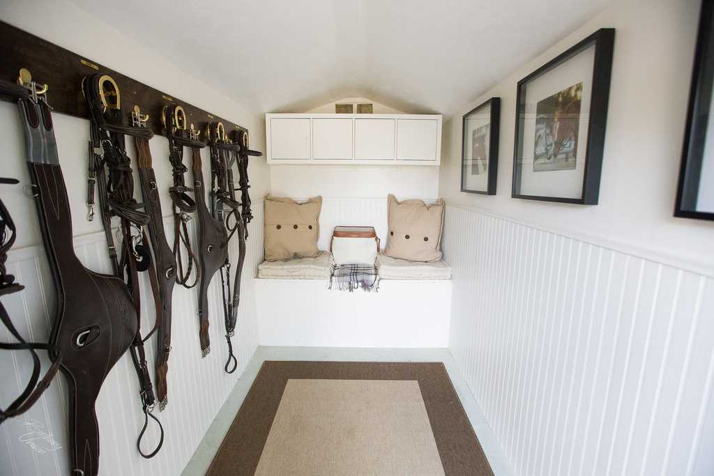 Equestrian tack room with saddles and bridles on the left, cushioned bench and framed photos on the right, and white cabinets above.