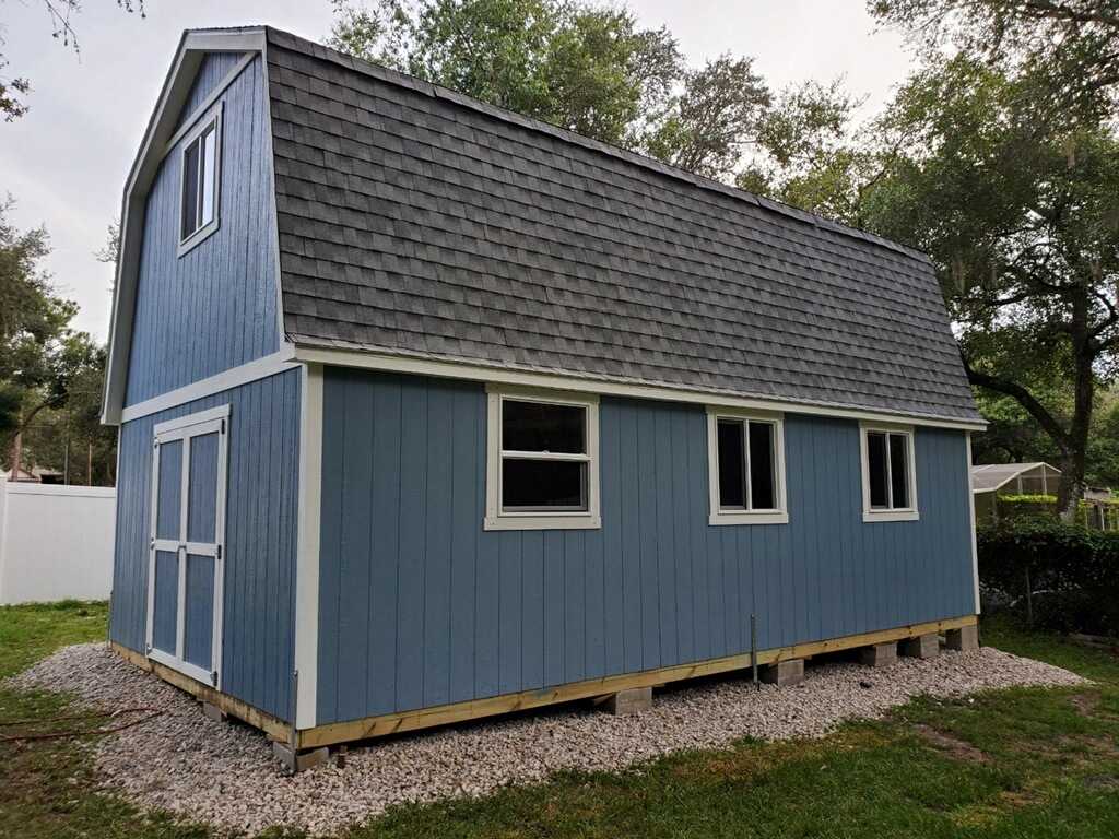 Front View of Gordon TB800 2S A blue two-story TB800 shed with a gray roof, white trim, and multiple windows, set on a gravel foundation surrounded by trees.