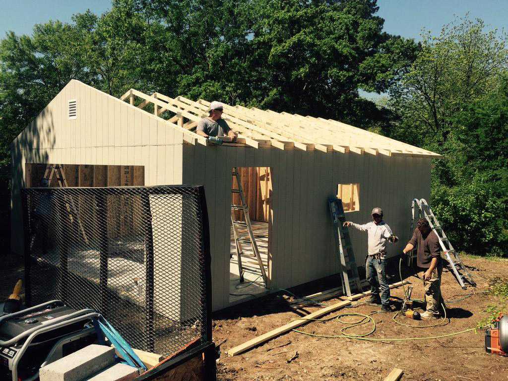 Sundance ranch garage under construction Two workers construct a Sundance ranch garage building with a wooden frame and partially completed roof, surrounded by trees and construction tools.