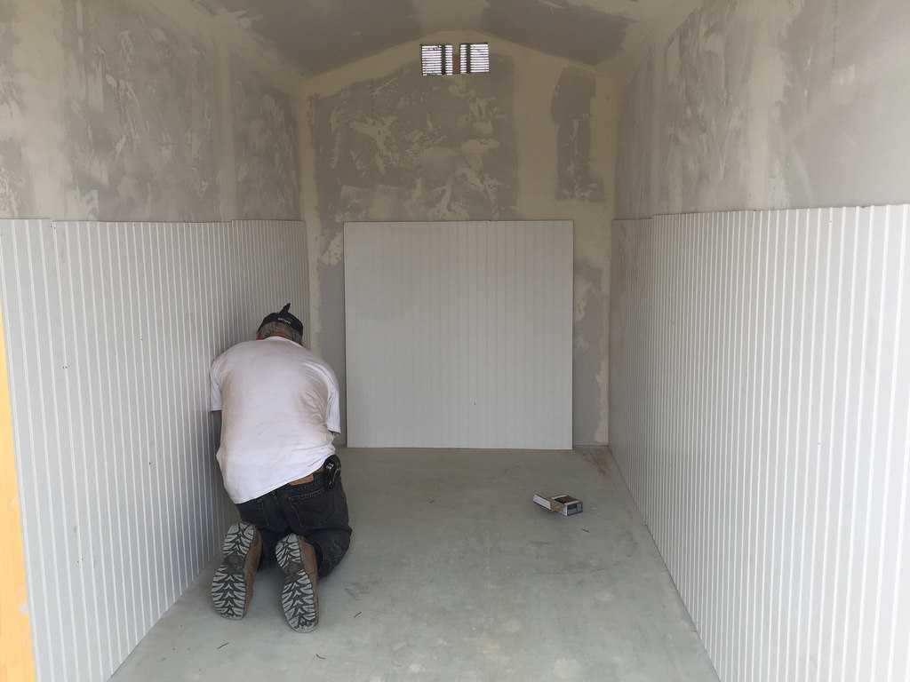 A man installing white paneling on the interior walls of a small, unfinished room with a concrete floor.