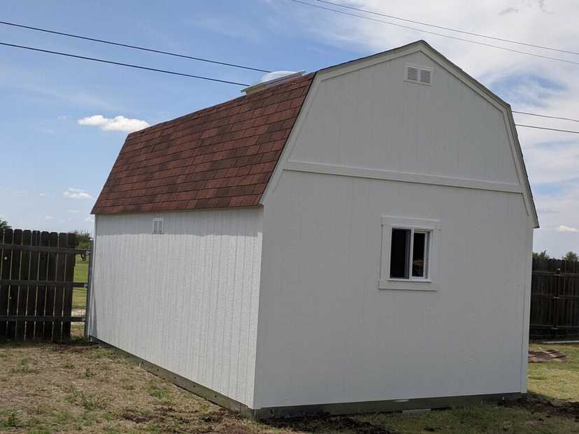 A white barn-style TB700 shed with a brown shingled roof, small window, and fenced yard under a partly cloudy sky.