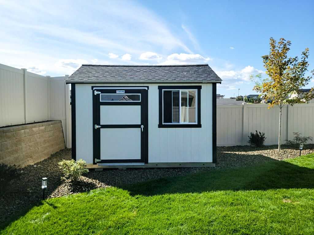 A TR700 white and black storage shed with a window and door, set in a fenced backyard with green grass and a young tree.