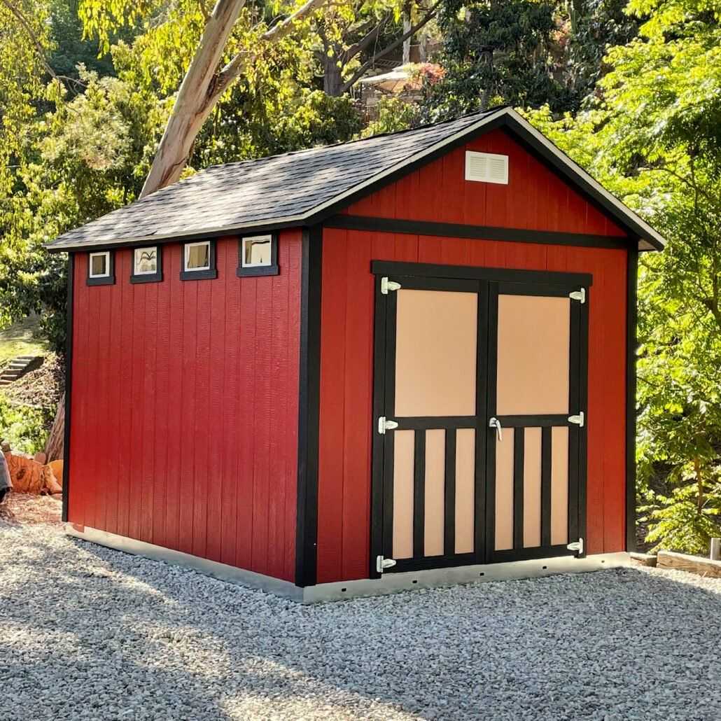 A red shed with black trim and a gable roof, surrounded by trees, sits on a gravel area. Three small windows are on one side.