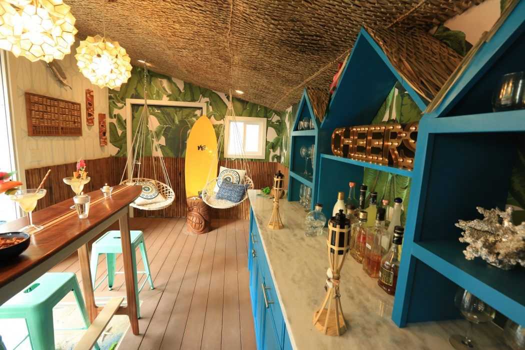 Tropical-themed bar with surfboards, hanging chairs, blue shelves, and a wooden table with stools, decorated with plants and lights