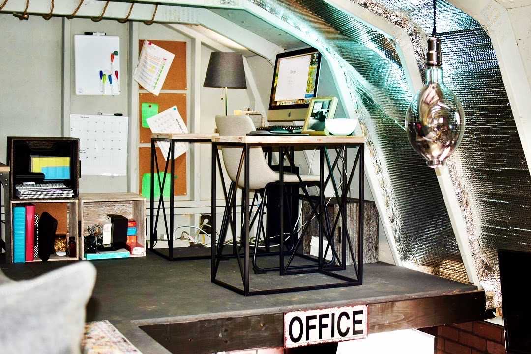 Modern home office in an attic with a desk, computer, lamp, and shelves. Insulated walls and a decorative bulb enhance the cozy atmosphere.