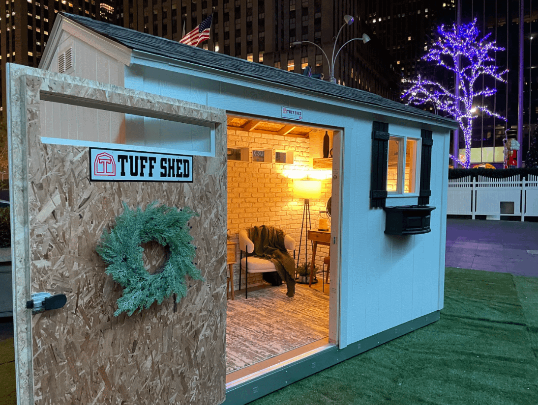 A small, illuminated shed with an open door, revealing a cozy interior. A wreath decorates the door, and a lit tree is visible outside.