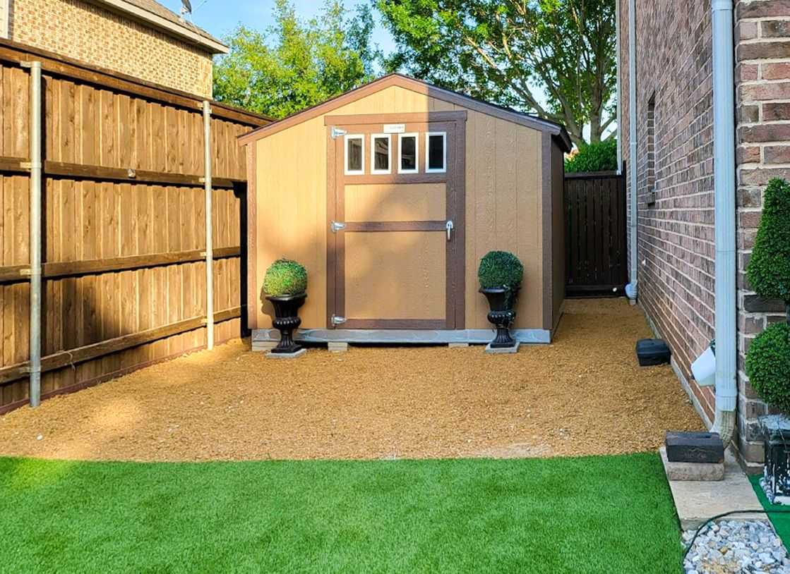 A SR600 brown shed with a white trimmed windows, flanked by two potted topiary plants, stands on a gravel area beside a brick wall.