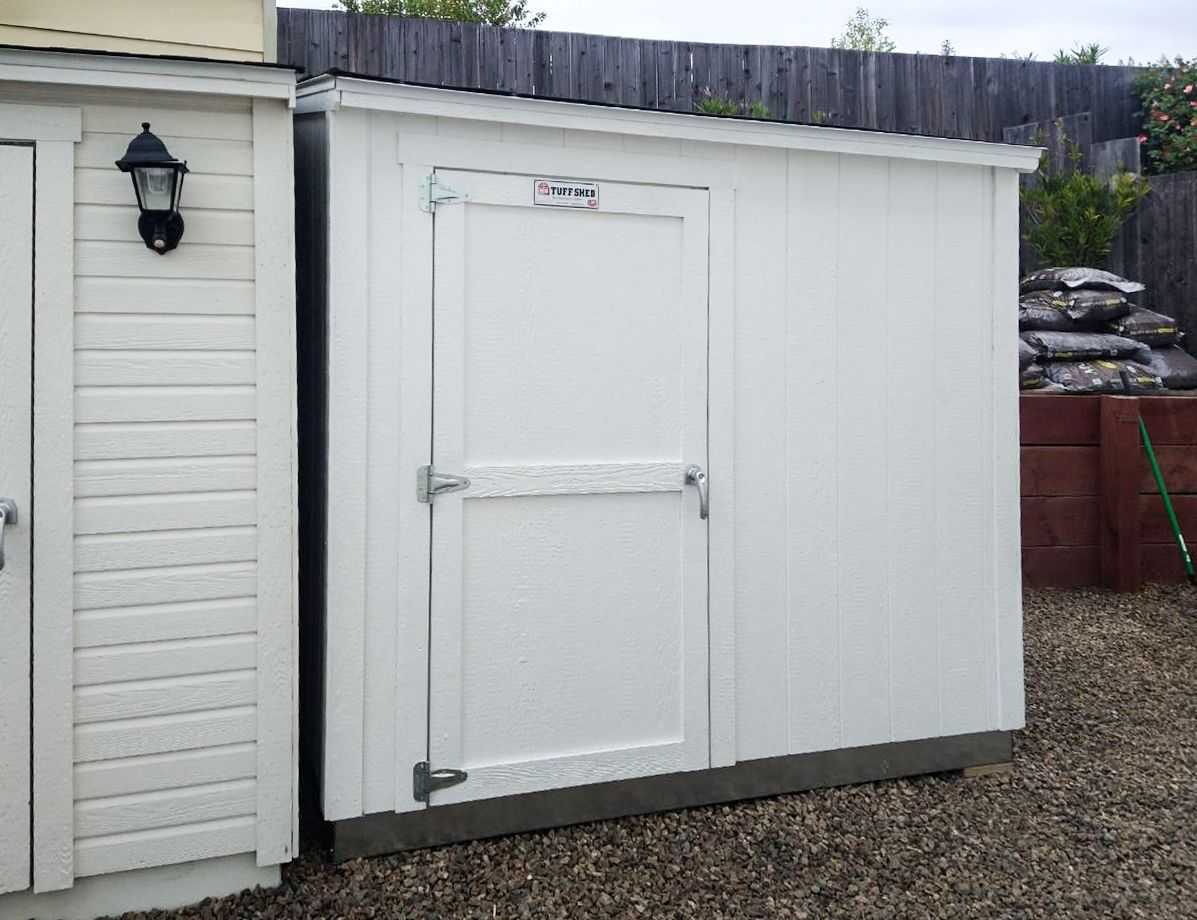 A white outdoor storage shed with a single door, located on a gravel surface.