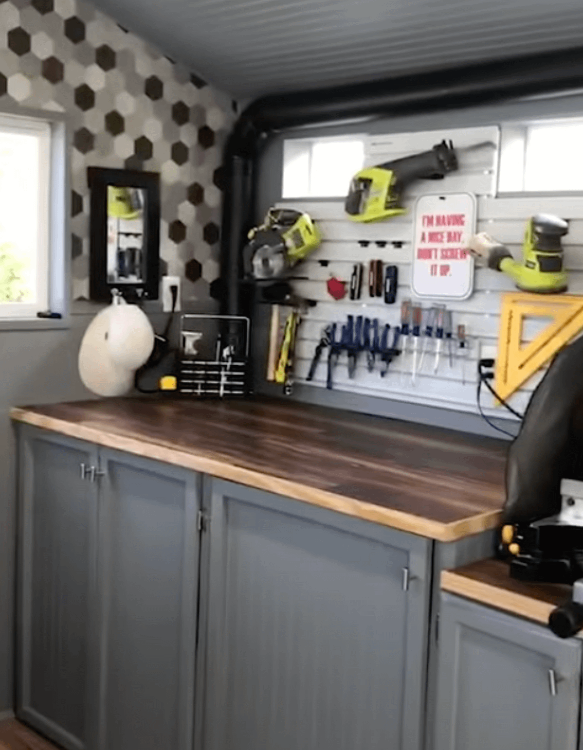 A tidy workshop with tools hanging on a pegboard above a wooden counter. Cabinets are below, and hexagonal tiles decorate one wall.