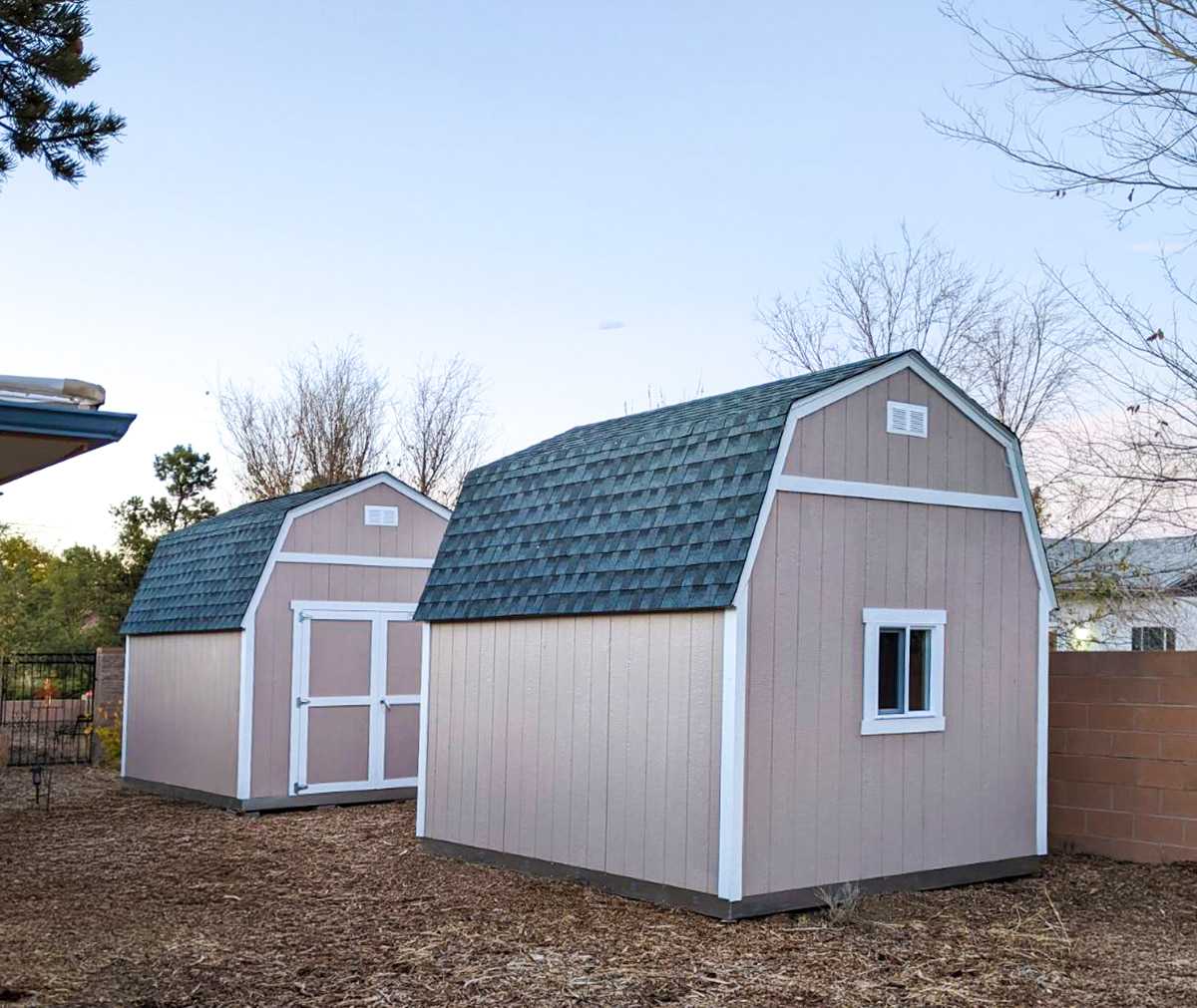 Two beige storage sheds with dark shingled roofs in a yard, surrounded by trees and a brick wall.