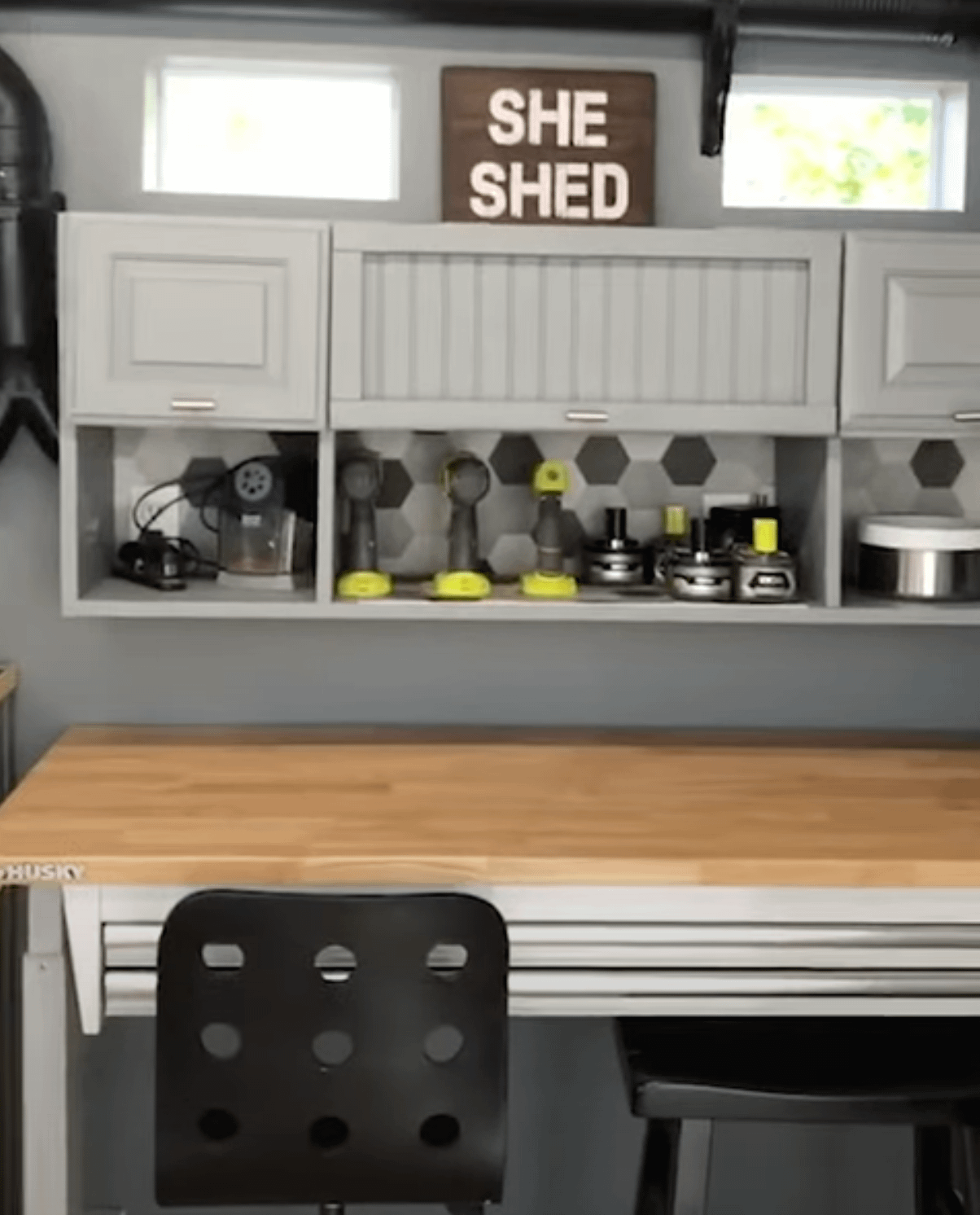 A well-organized workshop with a wooden countertop, black chairs, wall-mounted cabinets, tools, and a "She Shed" sign.