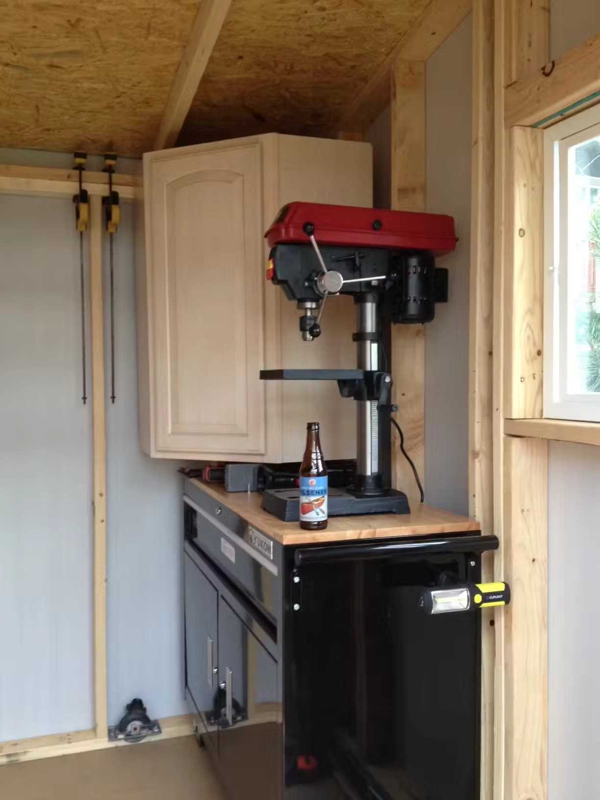 Inside a workshop, a red drill press is mounted on a black cabinet. A beer bottle rests in front. Clamps hang on the wall, and light enters through a window.