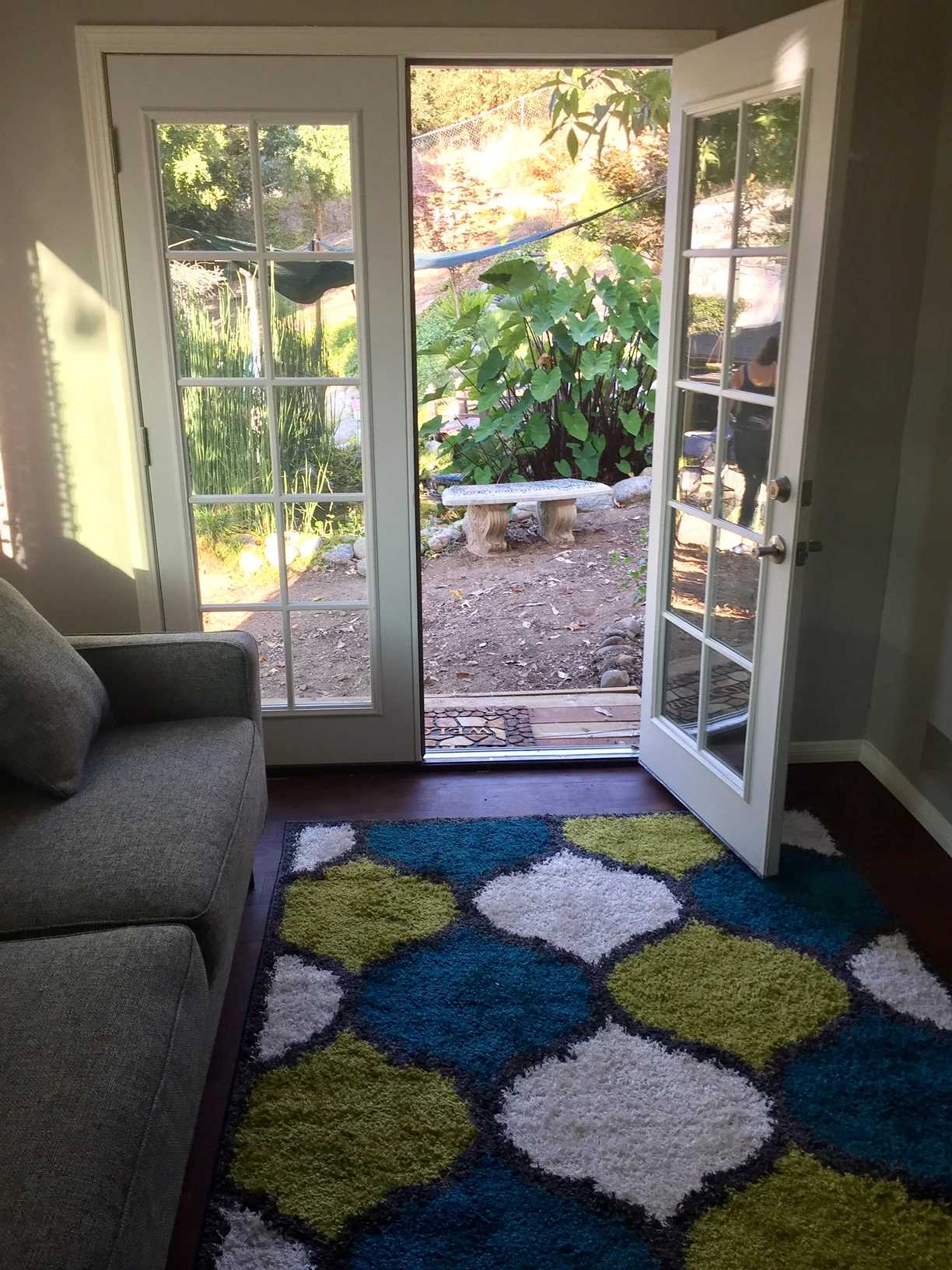 Open French doors lead to a garden with a wooden bench. Inside, a colorful rug and a gray sofa.