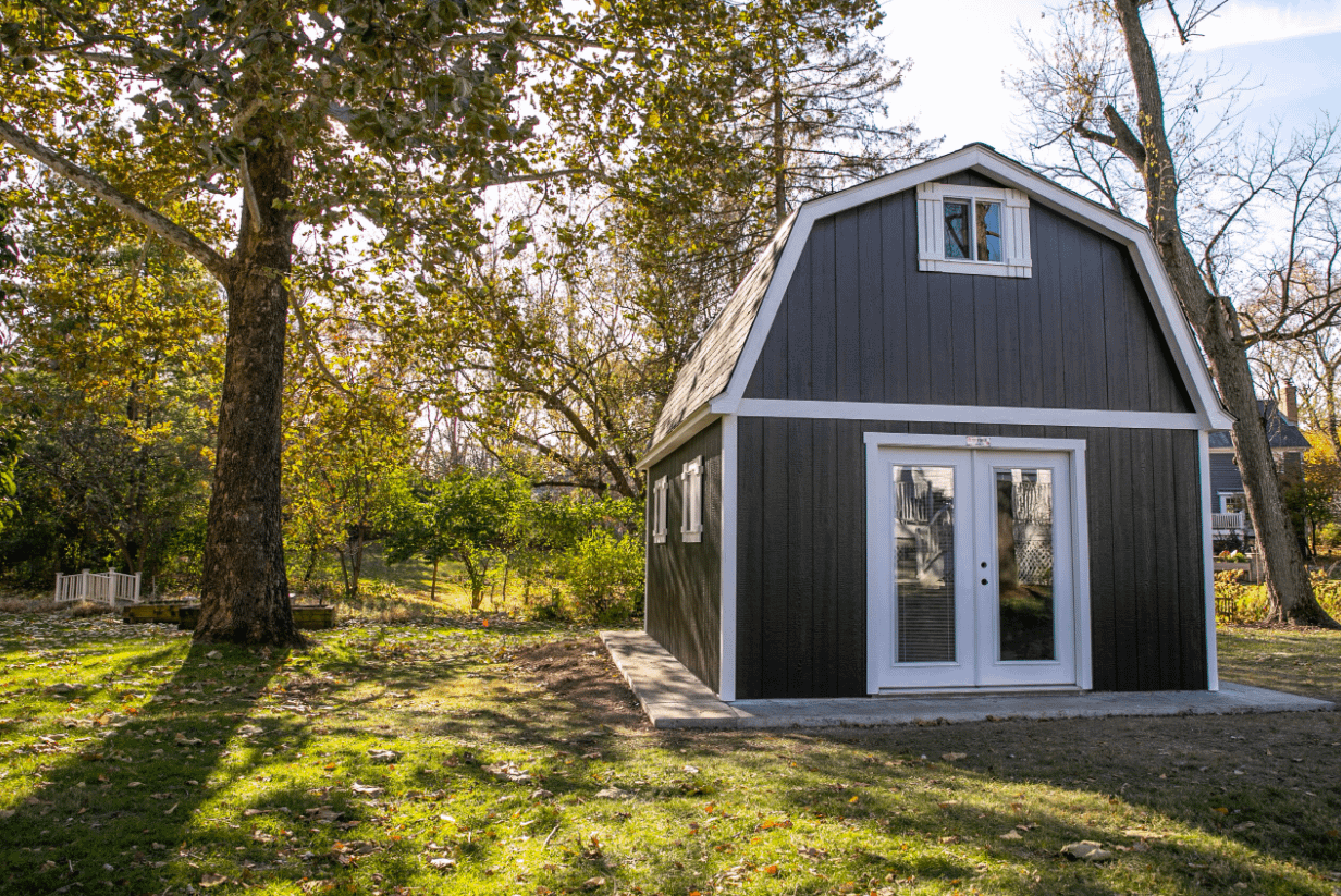 40th Anniversary Tuff Shed Building Example 1 A small, dark gray barn-style shed with double glass doors, surrounded by trees and grass, under a clear sky.
