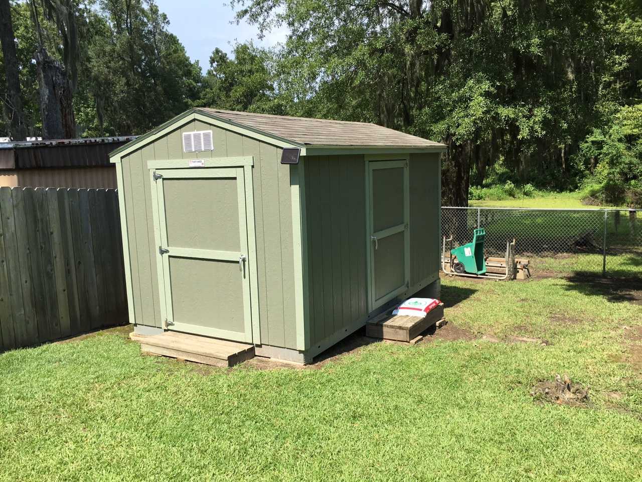 SR600 Exterior Shed A small green shed with a double door stands on a grassy lawn, surrounded by a wooden fence and trees in the background.