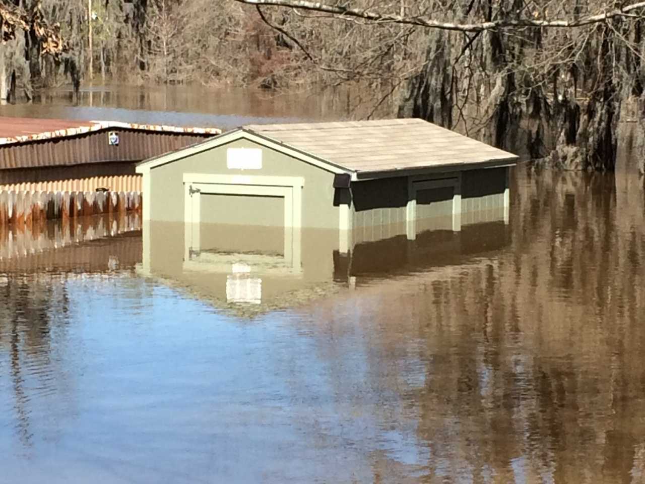 SR600 Submerged in Floodwater 1 A small green building partially submerged in floodwaters, surrounded by trees and debris, with water reflecting its structure.