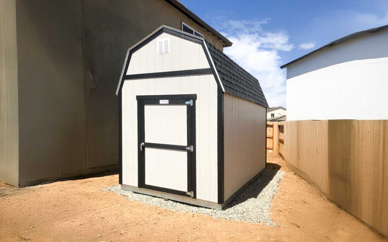 A beige shed with a black roof and trim stands on a gravel base between two buildings under a clear blue sky.