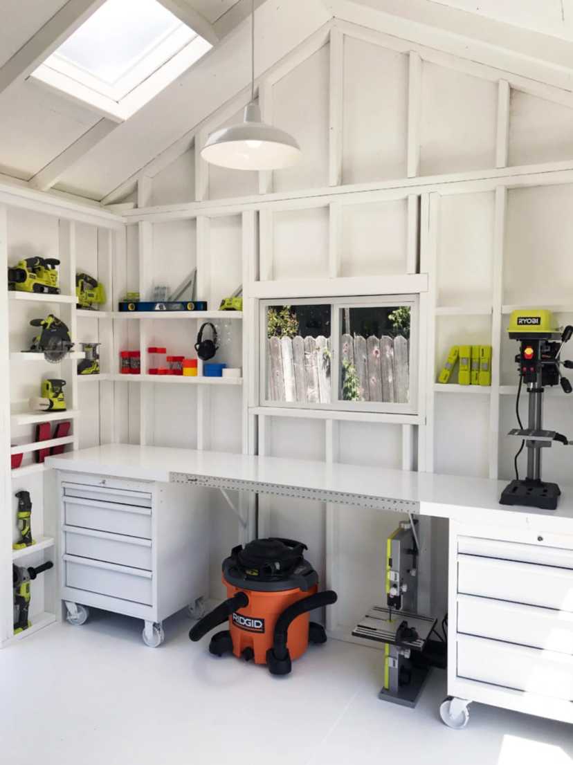 Brightly lit workshop with white shelving, workbenches, and various power tools. An orange vacuum is on the floor; a window shows a garden fence.