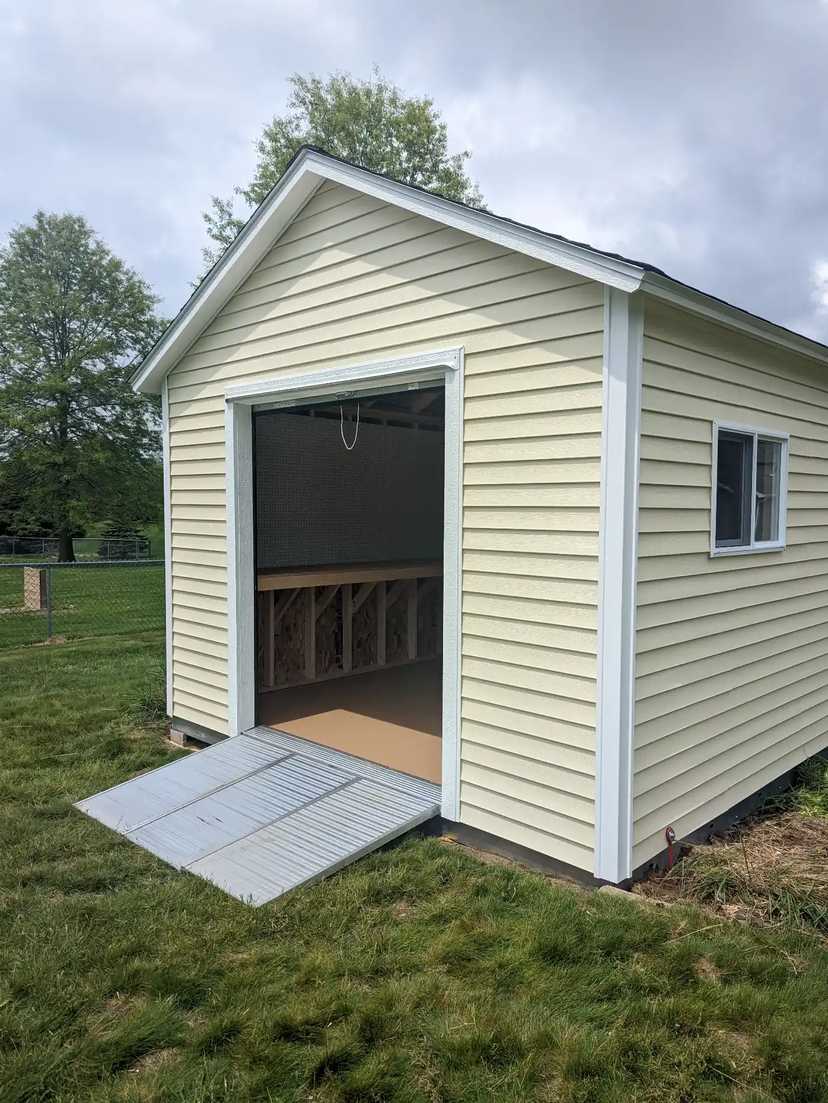 A small beige storage shed with a metal ramp, white trim, and a window, set in a grassy yard with trees in the background.