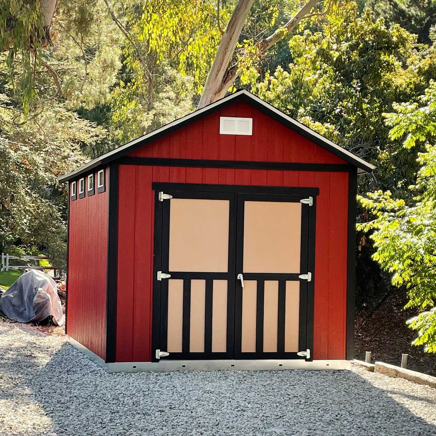 A small red and beige shed with black trim, surrounded by trees and gravel, under a clear sky.