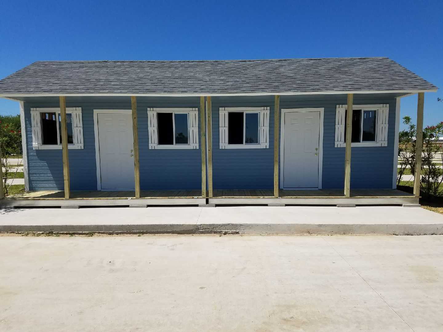 A small blue building with a gray roof, featuring three doors and windows, stands on a concrete surface against a clear blue sky.