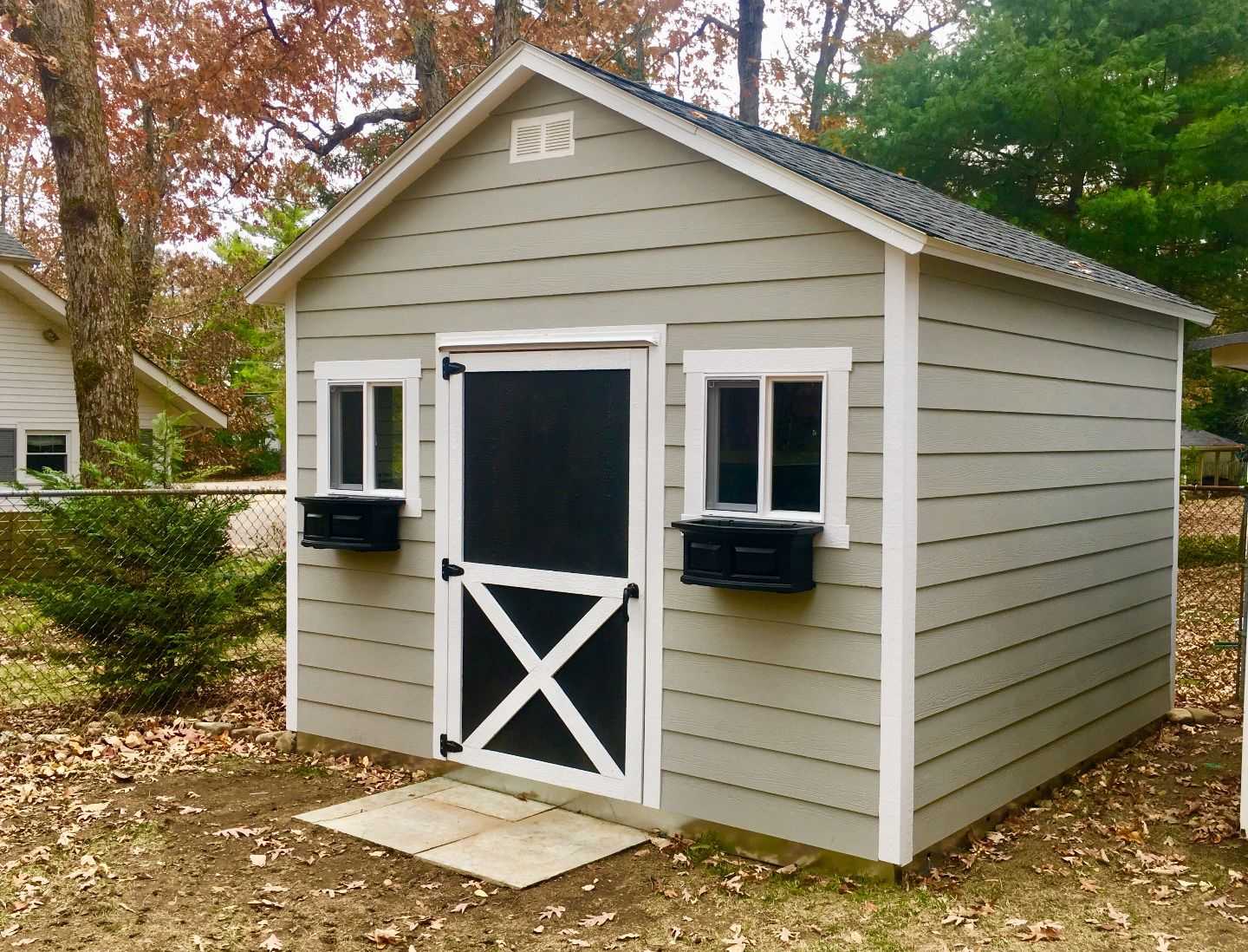A small, gray garden shed with a black and white door, two windows, and attached black flower boxes, surrounded by autumn leaves.