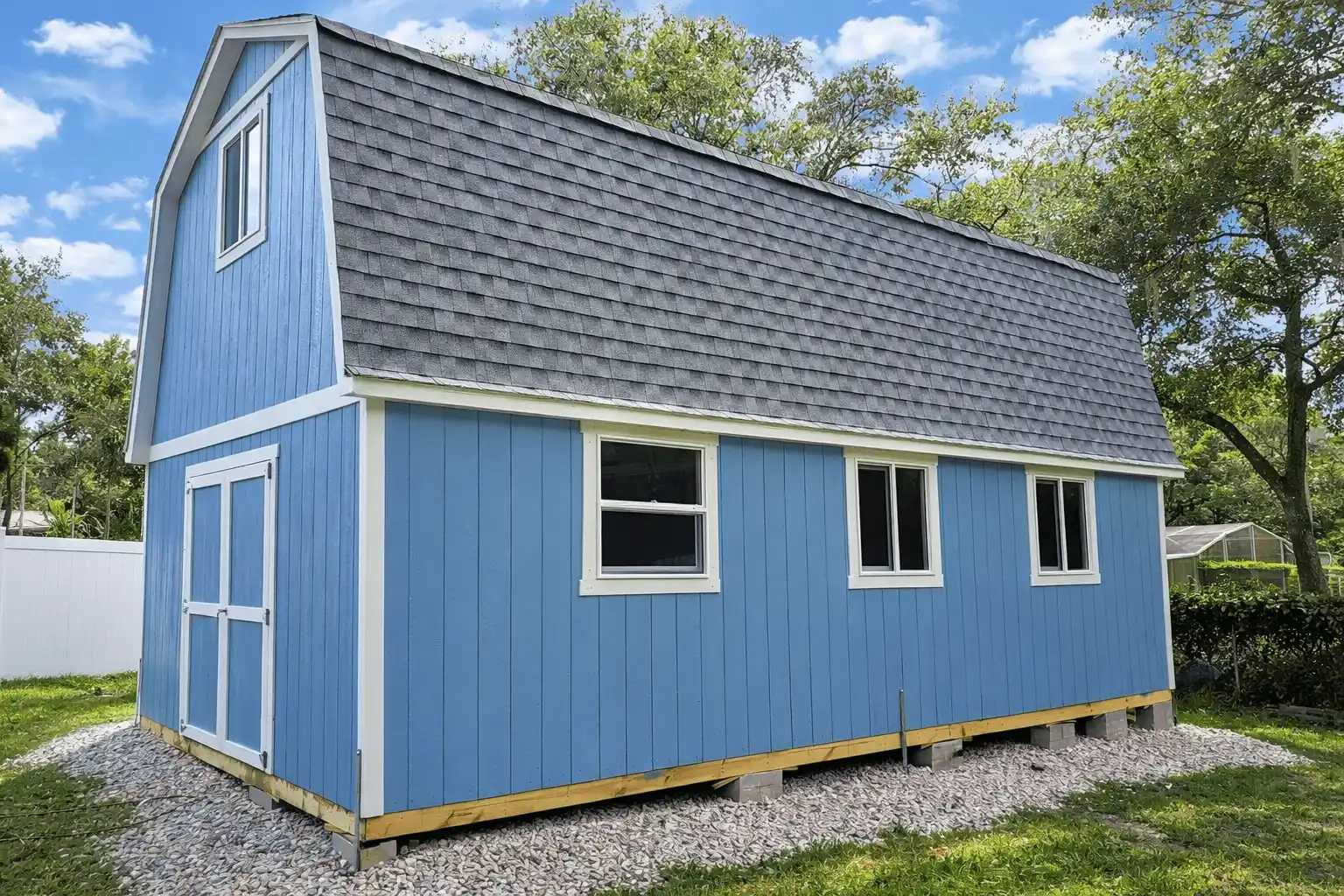 A blue barn-style shed with a gray shingled roof, white trim, and three windows, set on a gravel foundation surrounded by greenery.