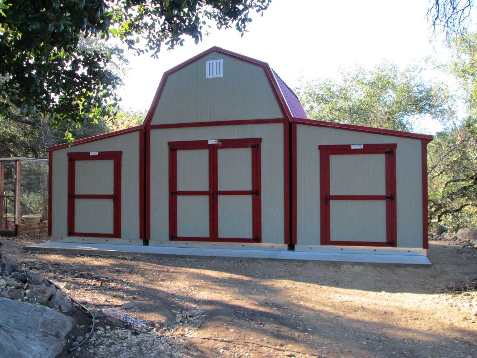 A large multiple beige and red barn-style shed with three doors, set on a concrete slab, surrounded by trees