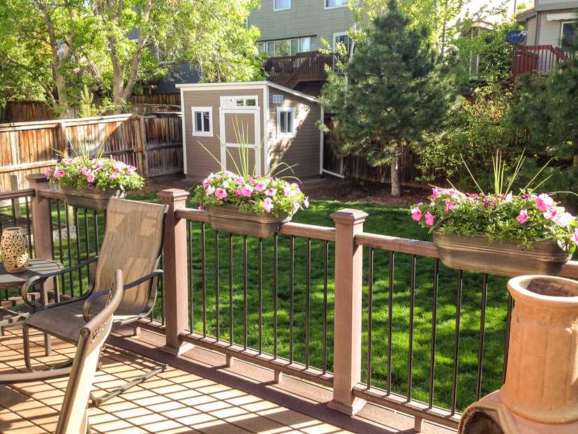 A sunny backyard with a wooden deck, potted flowers on the railing, a lawn, and a Sundance Lean-To surrounded by trees and a wooden fence.