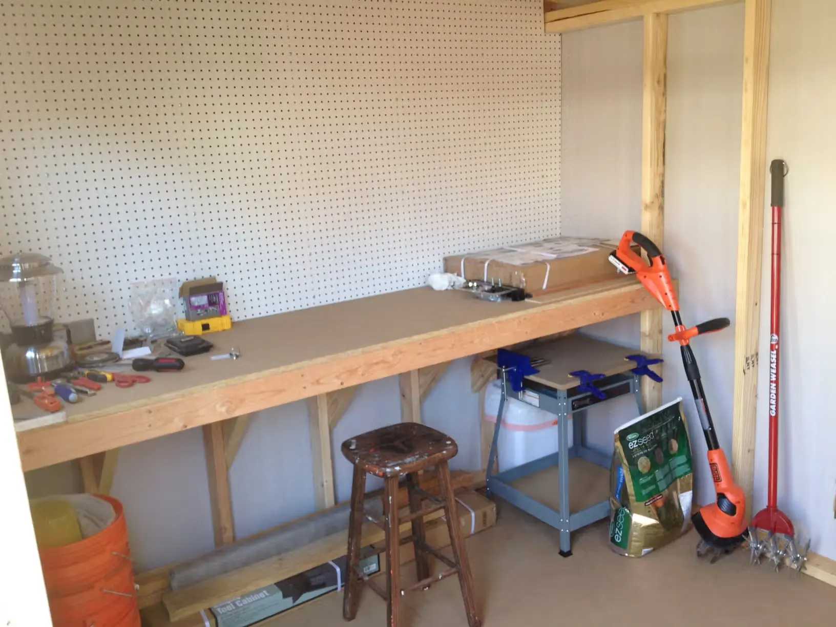 A tidy workshop with a wooden workbench, tools, and gardening equipment. A stool and storage below the bench, and a string trimmer leaning against the wall.