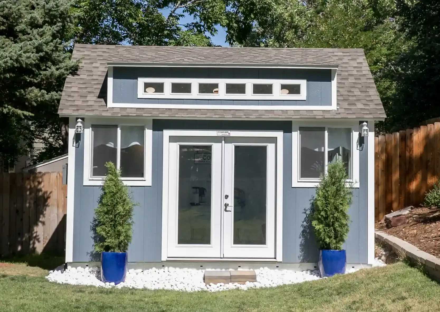 A small blue shed with white trim, double glass doors, and six narrow windows. Two potted plants in blue pots flank the entrance.