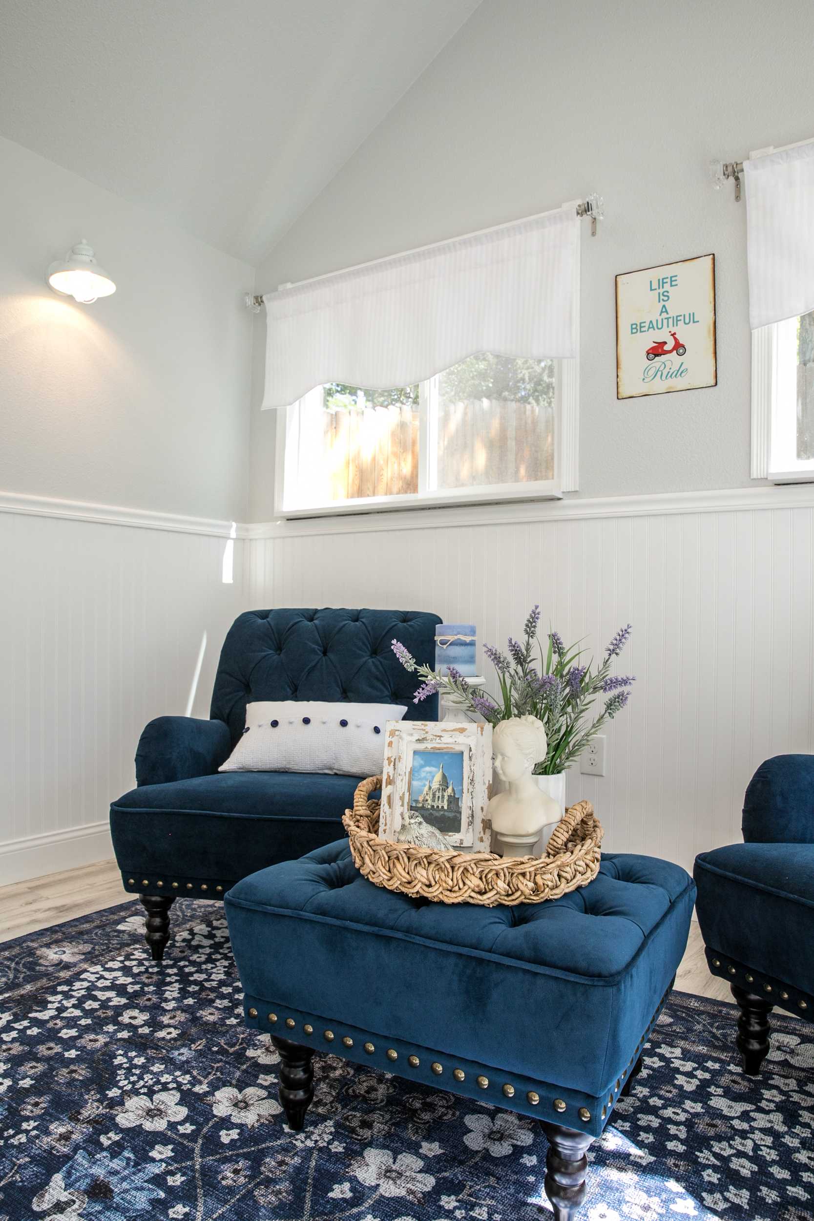 Linda's Interior TR-800 She Shed 3 Cozy room with blue velvet chairs, floral rug, and decorative tray on ottoman. Wall art and soft lighting create a serene atmosphere.
