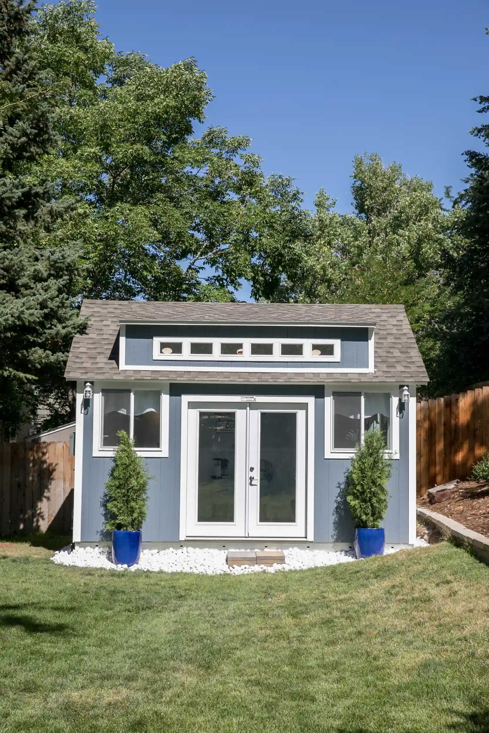 Small blue garden shed with white trim, flanked by two potted trees in blue pots. Surrounded by grass and trees under a clear blue sky.