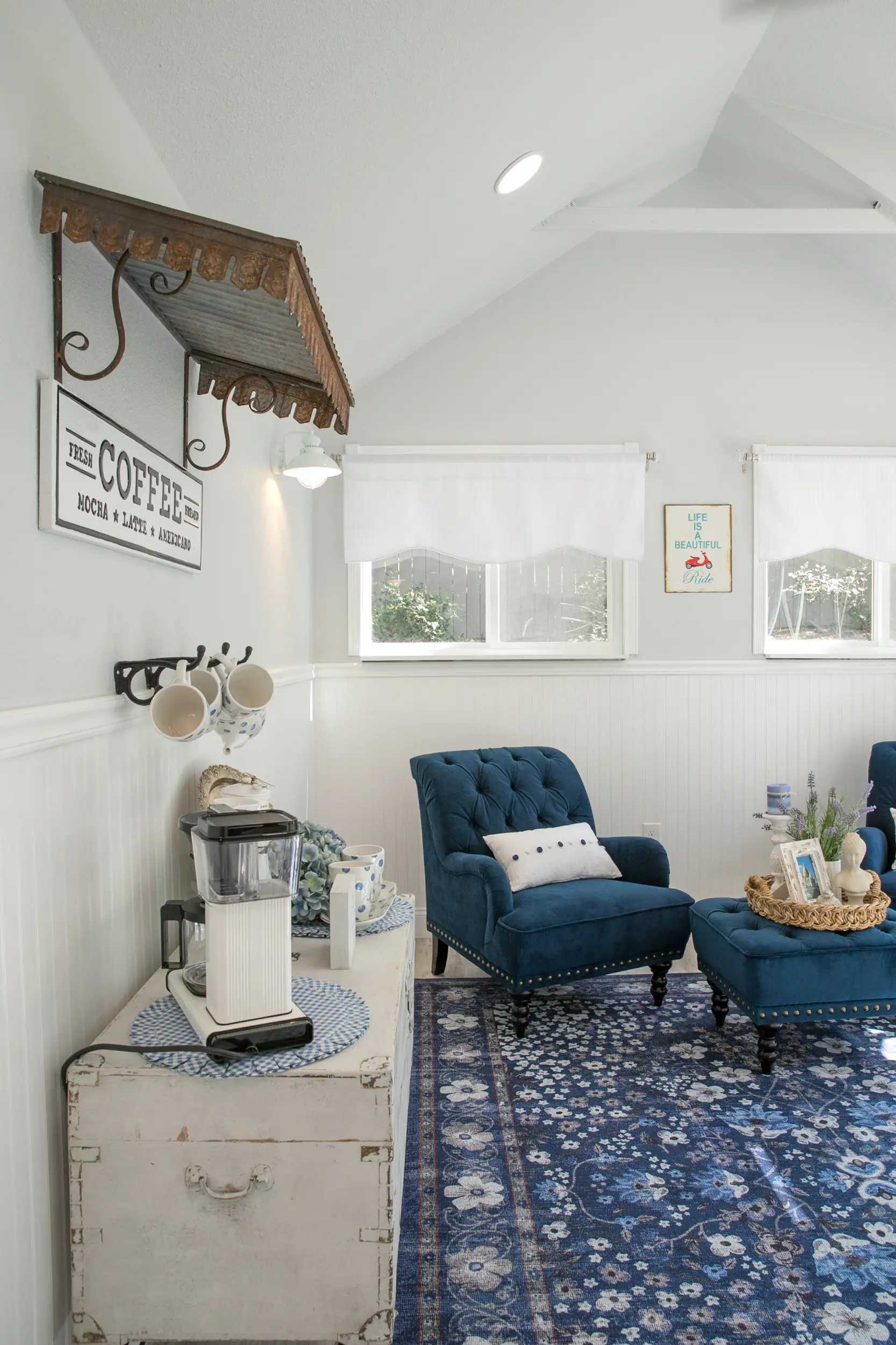 Cozy room with blue armchairs, patterned rug, vintage coffee station, and white walls under a sloped ceiling. Bright windows add natural light.