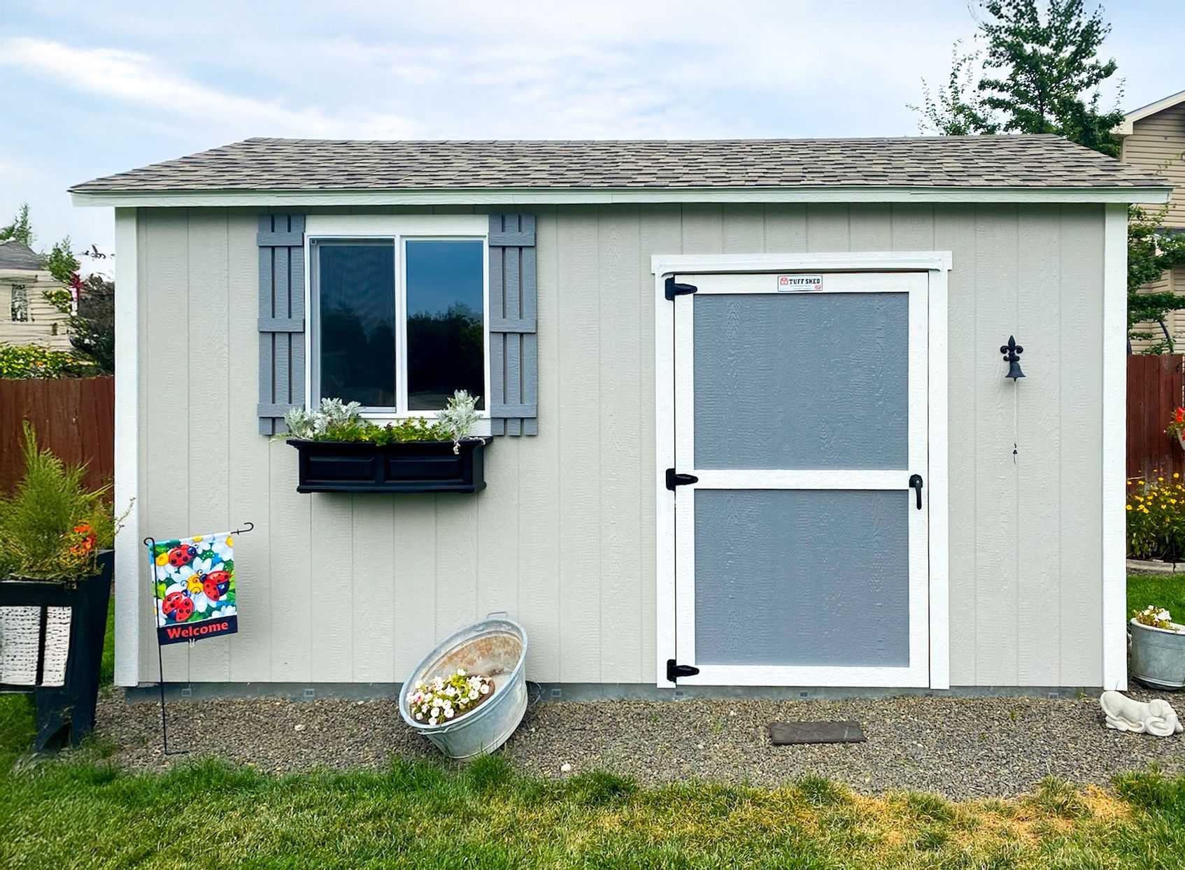 A TR800 gray shed with a window box of plants, a welcome flag, and a metal tub with flowers, set in a grassy backyard.