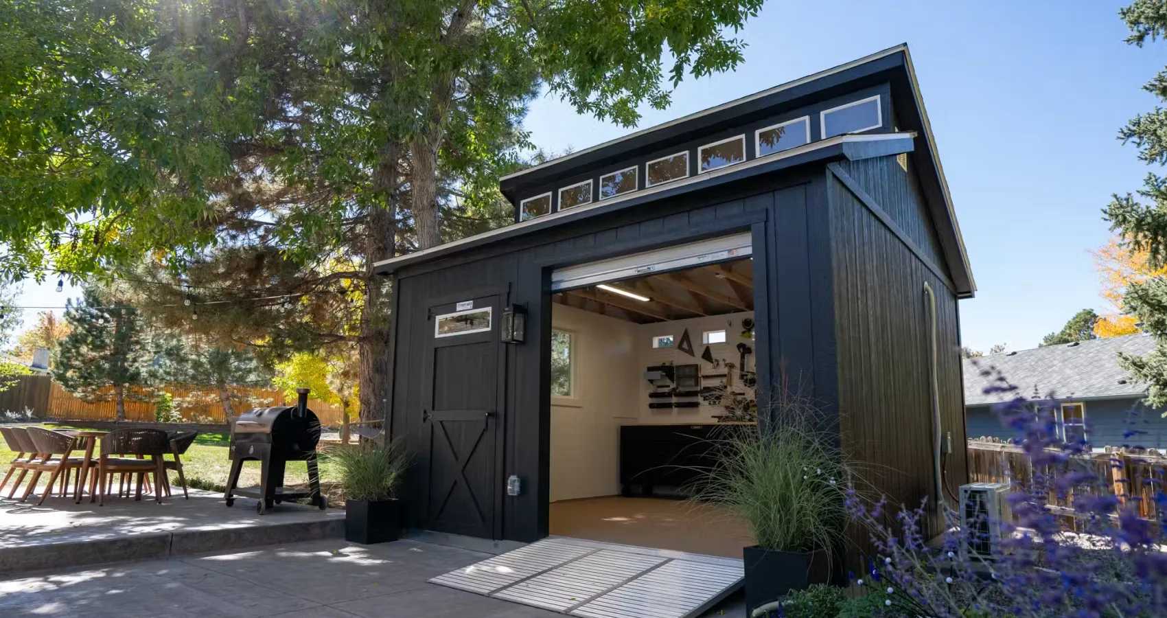 A modern black shed with open double doors, surrounded by trees and plants, under a clear blue sky. A barbecue grill is visible to the side.