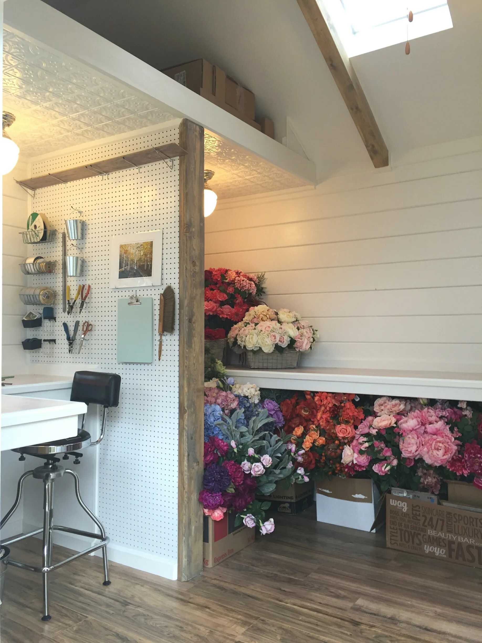 Cozy florist workspace with white walls, pegboard for tools, and vibrant flowers in baskets and boxes, lit by hanging lamps