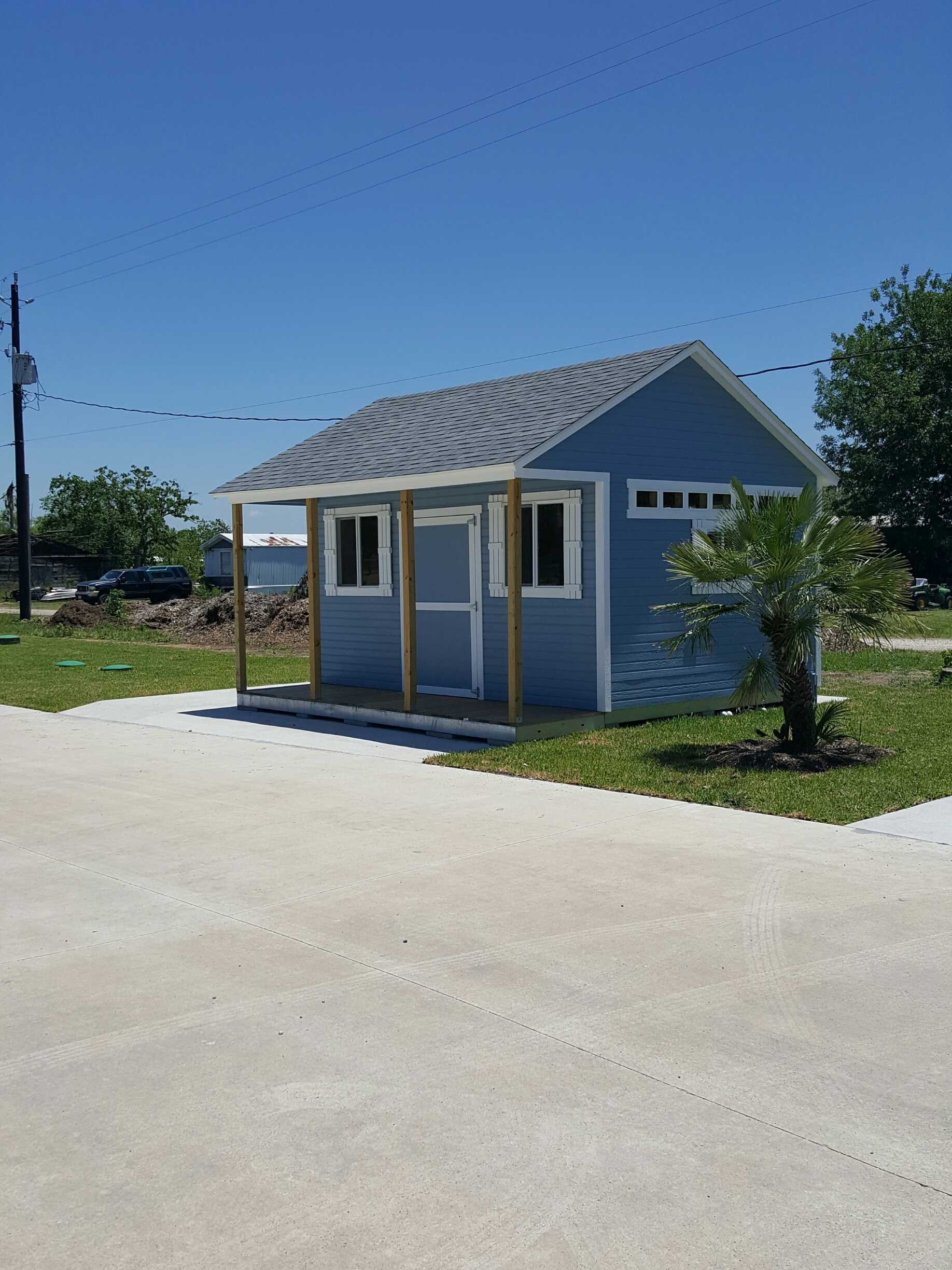 TR800 shed front with a gray roof and white trim, situated on a grassy area next to a palm tree, under a clear blue sky.
