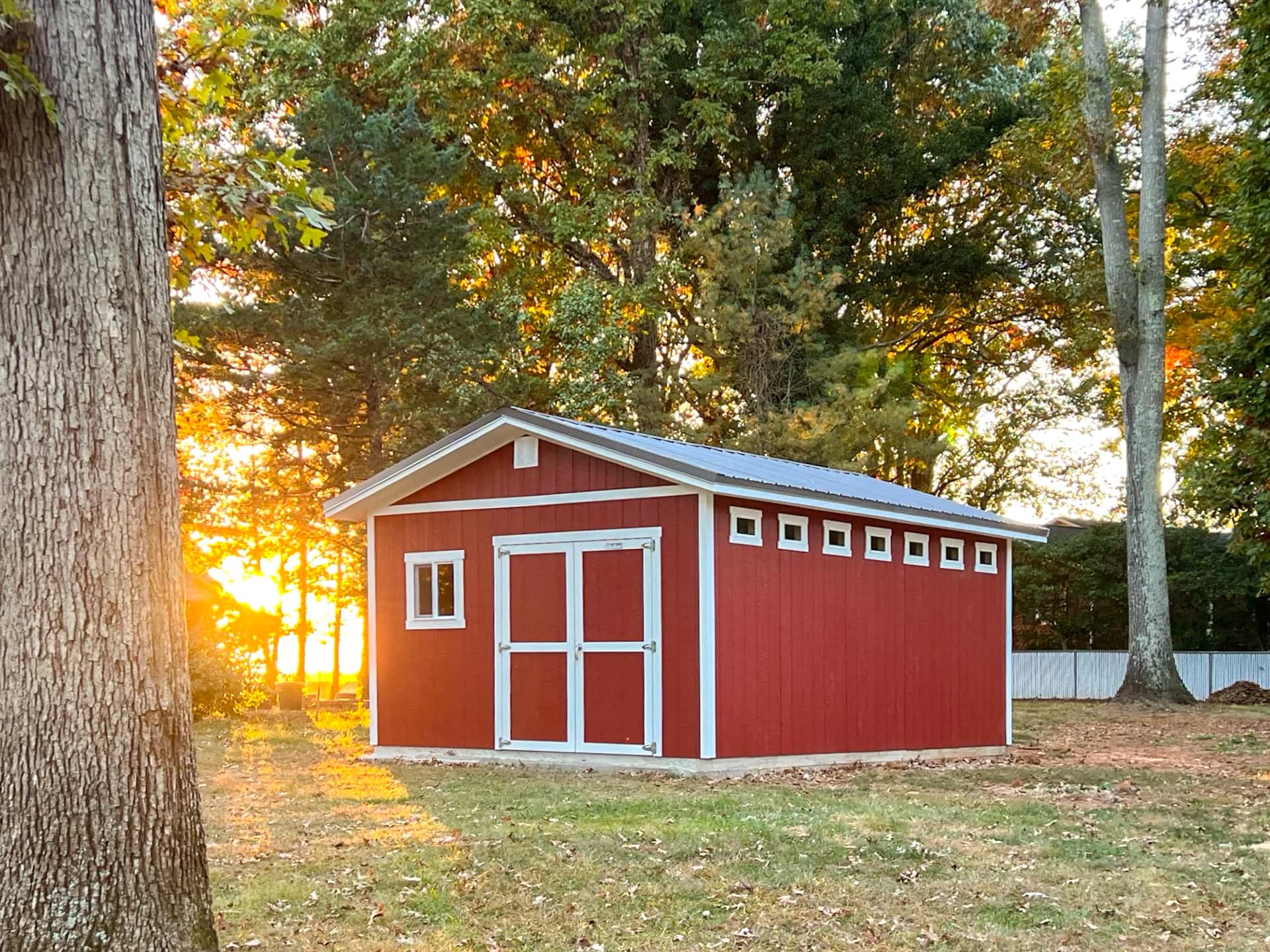 A TR800 red shed with white trim surrounded by trees, with the sun setting in the background.