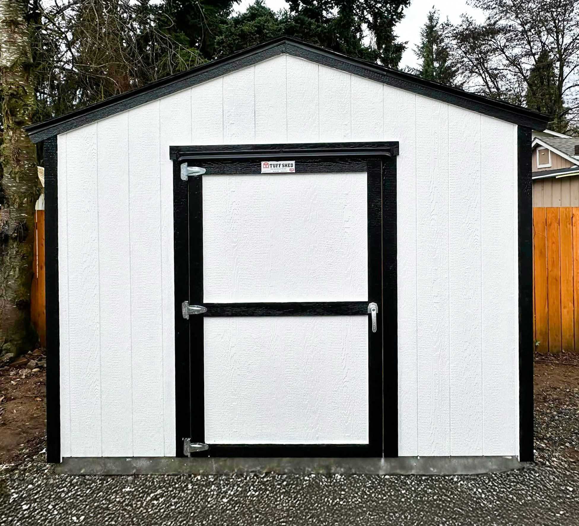 A white TR700 shed with a black-trimmed door and roof stands on a gravel surface.