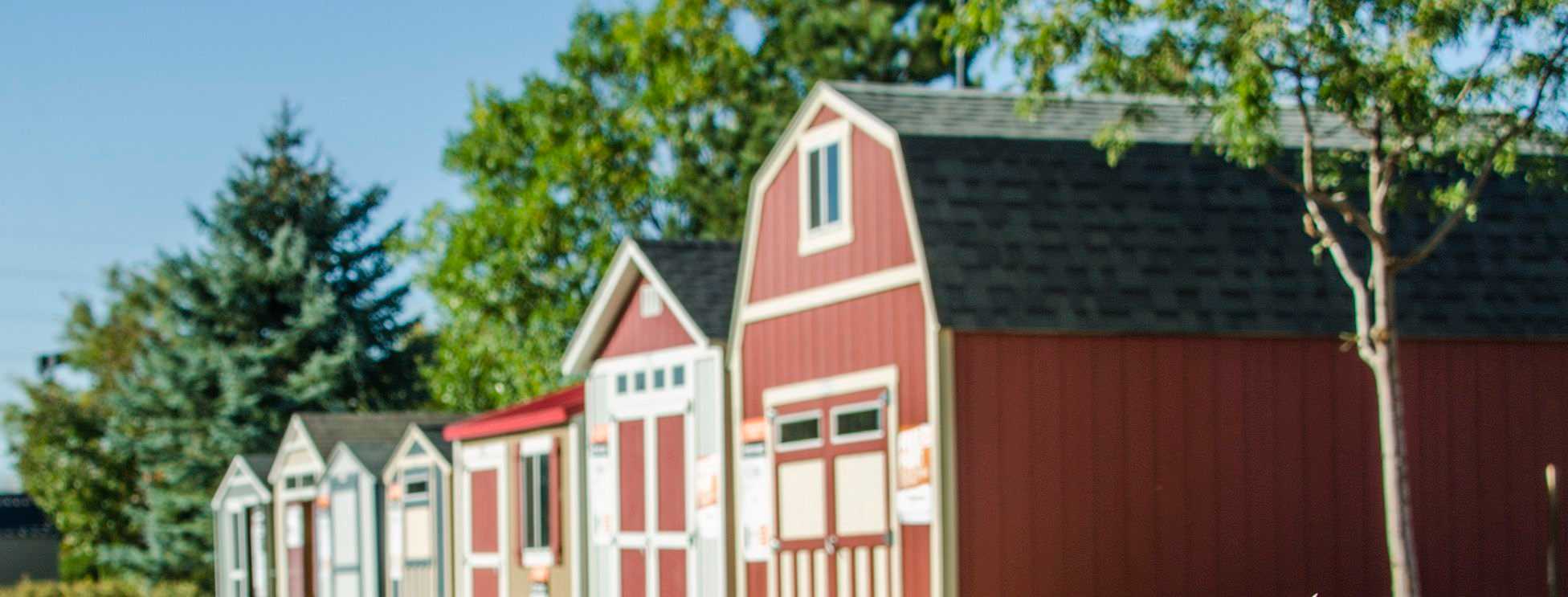 Row of red and white Tuff Shed barn-style sheds with trees in the background under a clear blue sky.