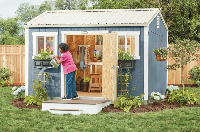 A woman in a pink shirt waters plants outside a small blue garden shed with an open door, surrounded by a neatly landscaped yard.