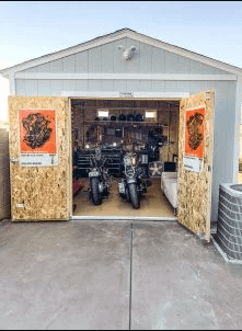 Two motorcycles parked inside an open garage with tools and equipment on shelves. Posters are on the doors, and a fan is visible inside.
