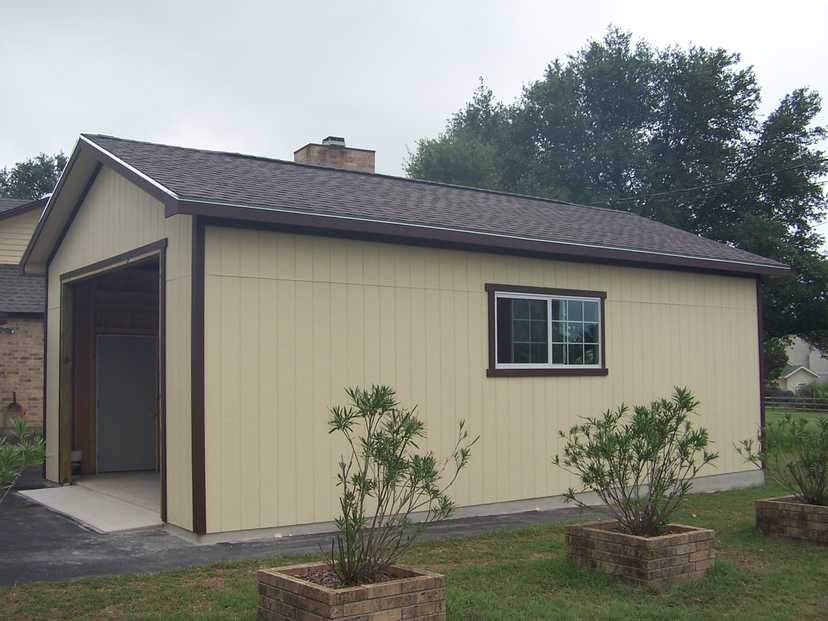 A beige detached garage with a brown trim, one window, and an open door, surrounded by trees and brick planters with small bushes.