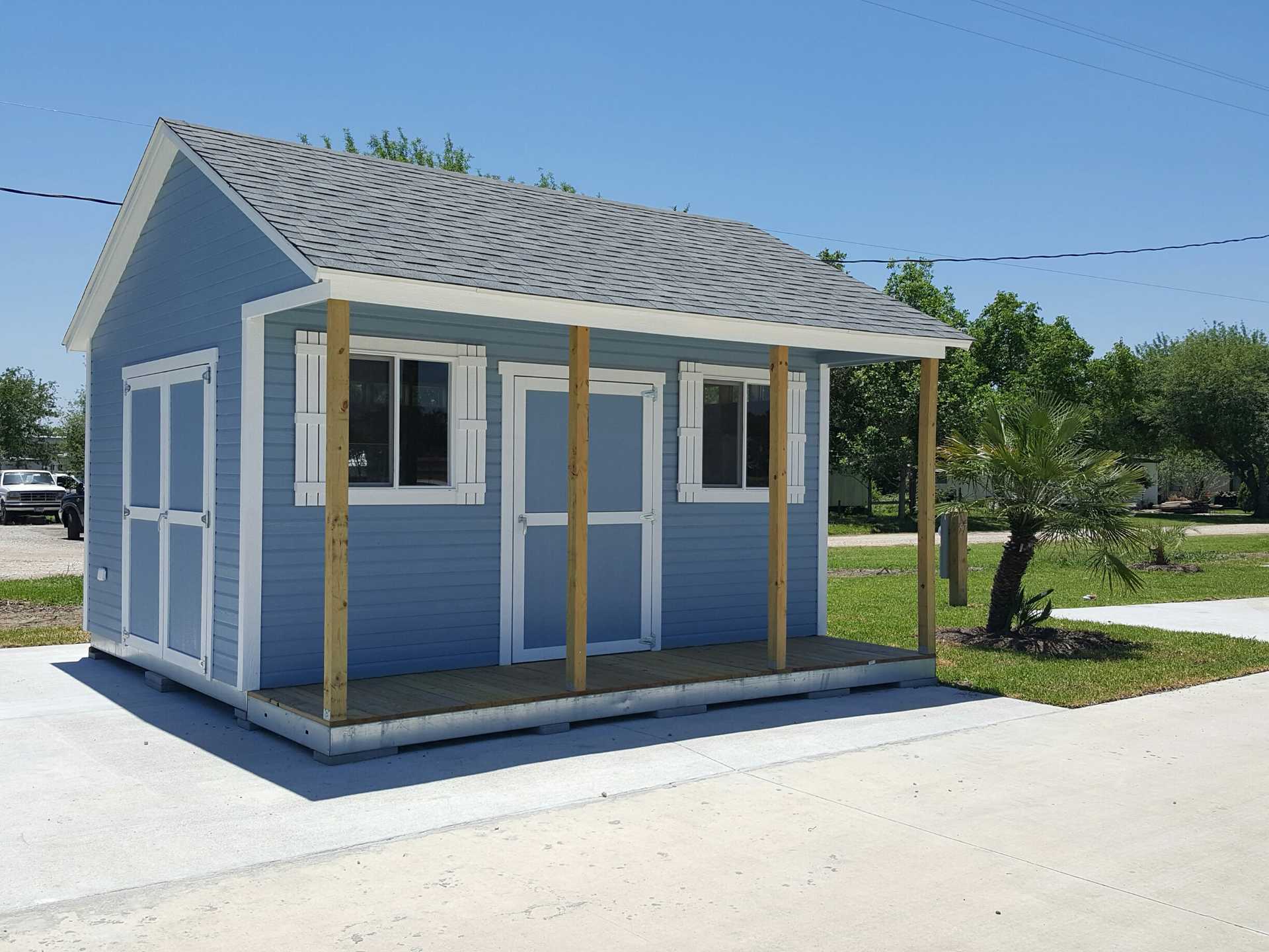 TR800 shed front with white shutters and a porch, set on a concrete slab, surrounded by grass and trees under a clear blue sky