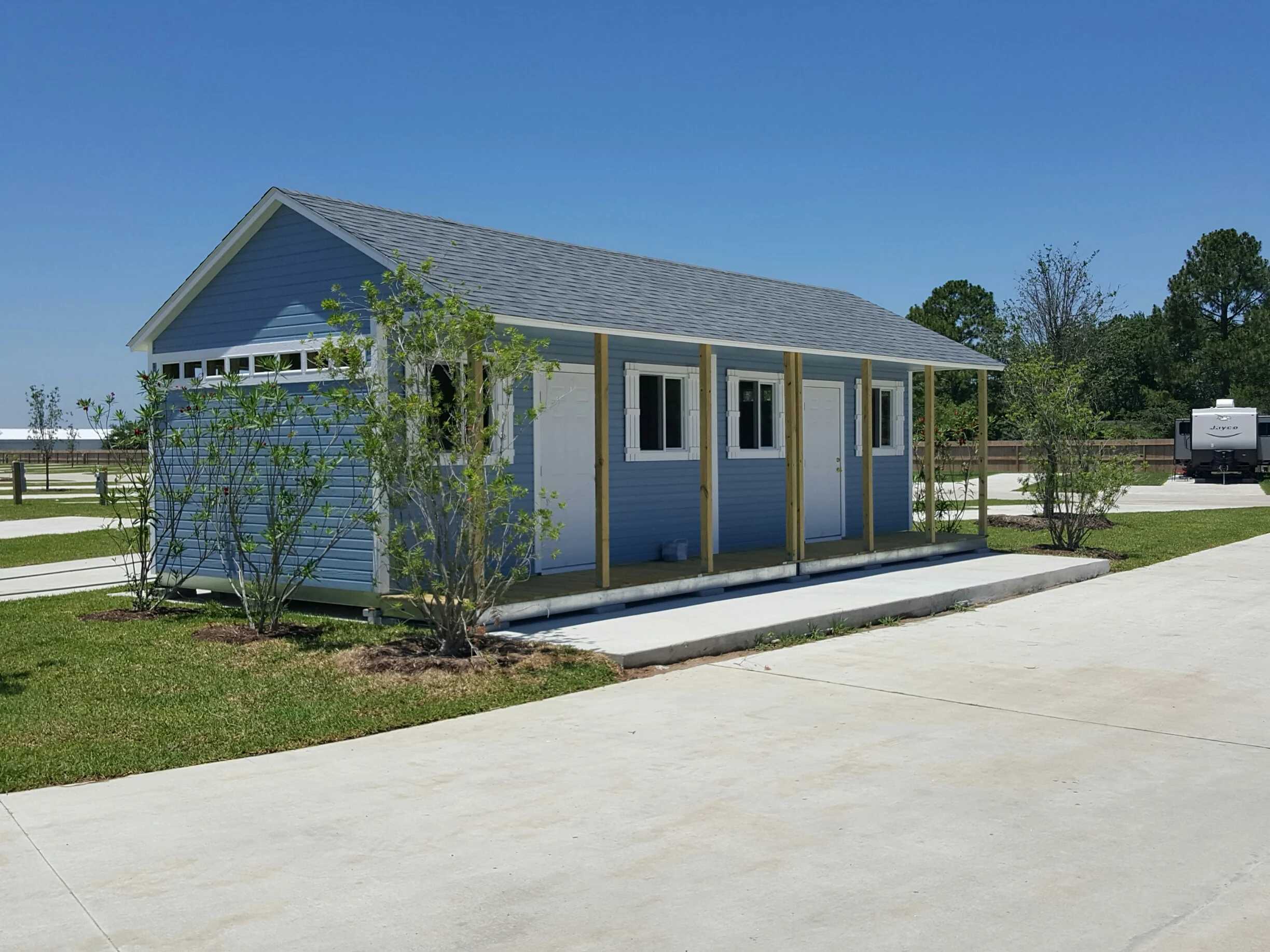 A small blue building with white trim and a gray roof, surrounded by grass and a few young trees, set against a clear blue sky.